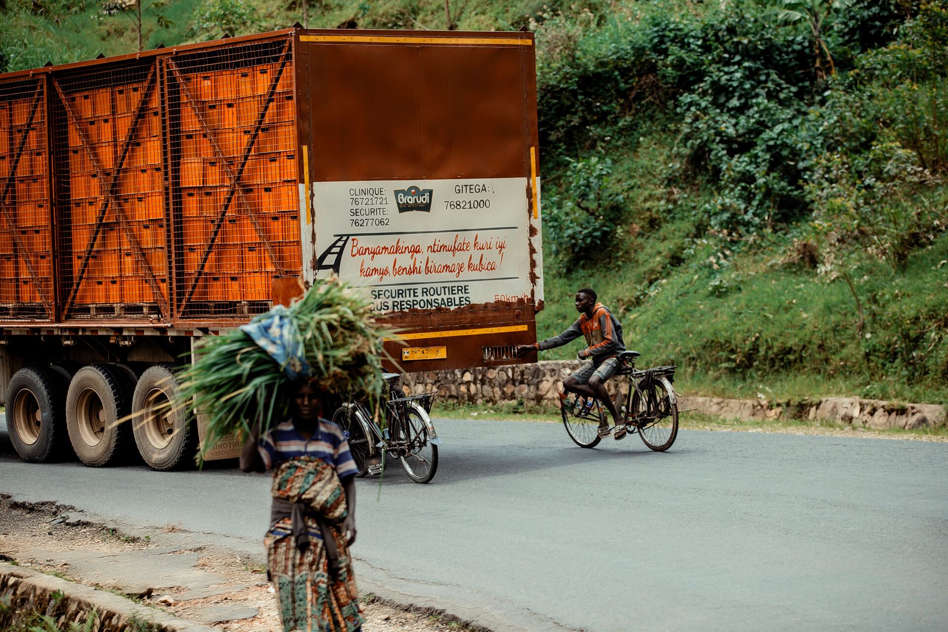 Cyclist holding on to a truck, rural road and a woman walking with a load, Congo DRC